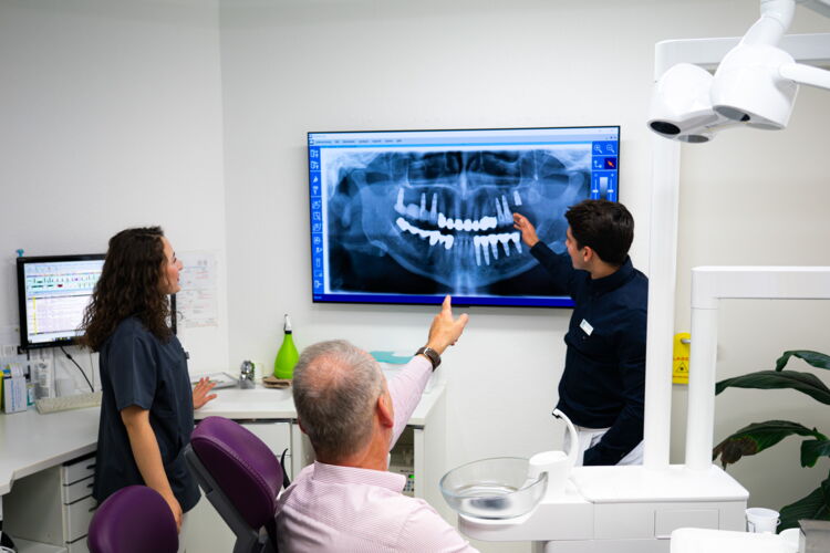 Zahnarzt, Helferin und Patient diskutieren auf Basis eines am Wandmonitor angezeigten Röntgenbild eines Gebisses